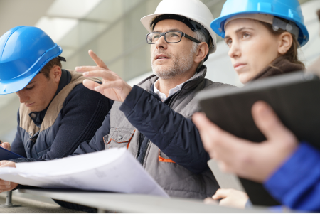 Image of two men and a woman in hard hats looking out at a project in the distance and holding a paper schematic
