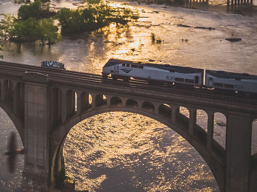 Image of an Amtrak train crossing the James River on the CSX A-Line Bridge in Richmond VA as the sun sets in the background and glows over the water