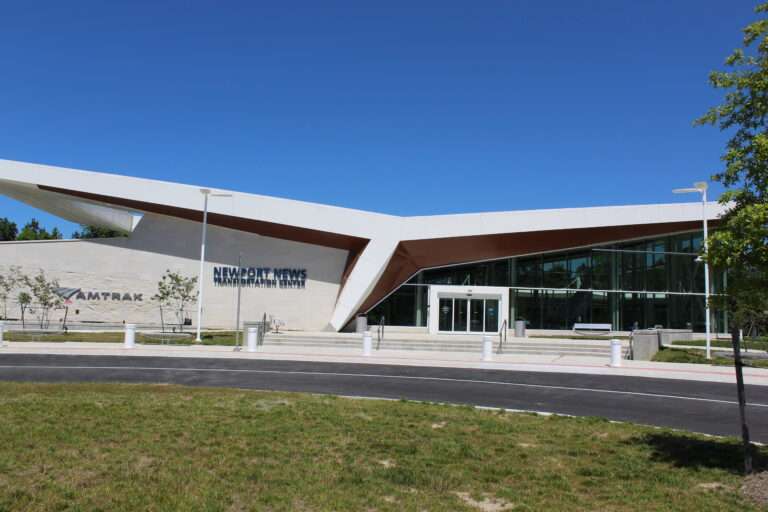 The Newport News Transportation Center, a modern Amtrak facility with an angular white and brown design, large glass windows, and a sleek entrance. A clear blue sky highlights its contemporary architecture