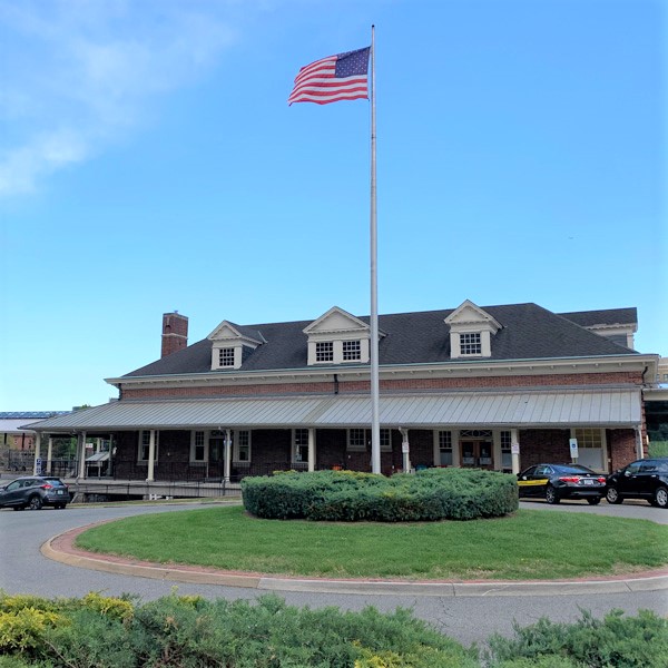 A historic train station with a flagpole featuring the American flag, surrounded by a circular driveway and greenery.