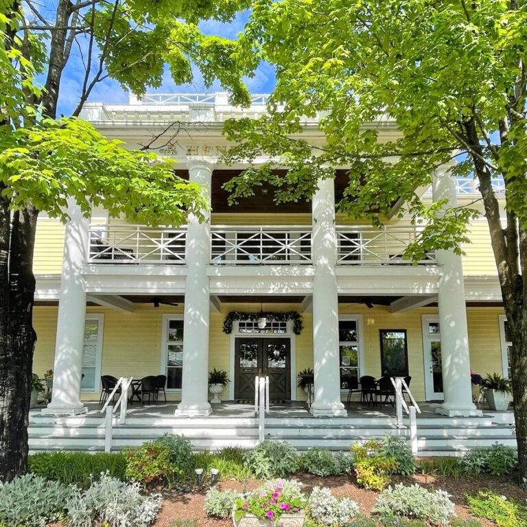 A large, two-story yellow building with white columns and a porch, surrounded by greenery and trees.